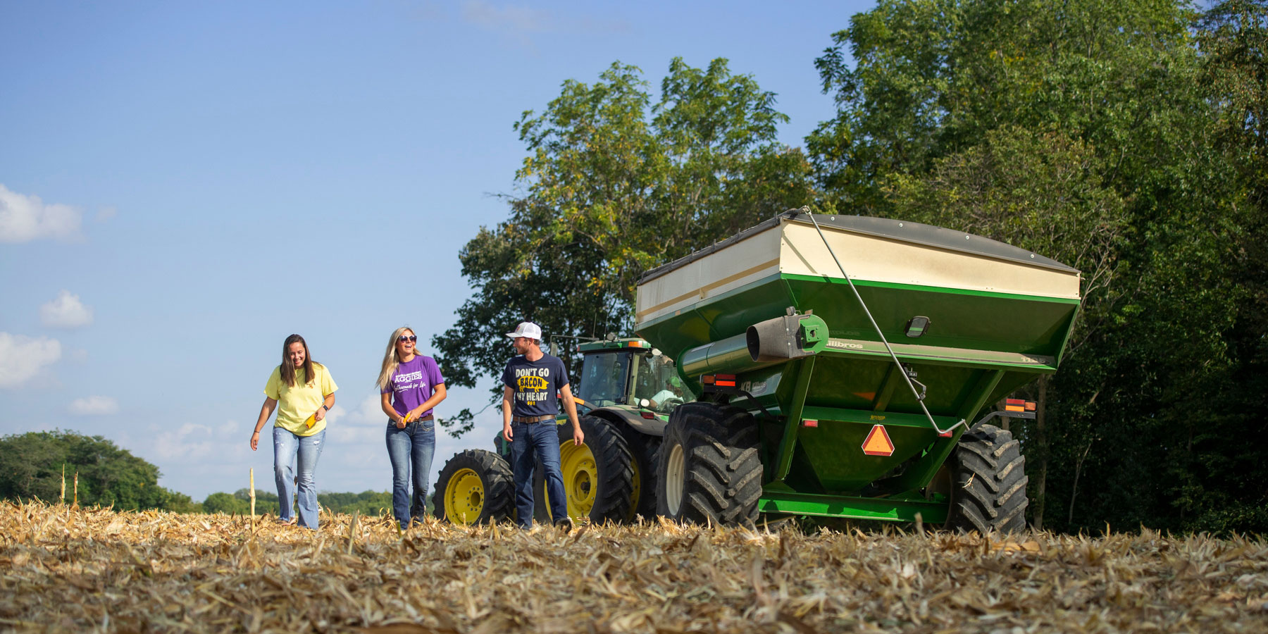 three students walking in harvested field next to harvester